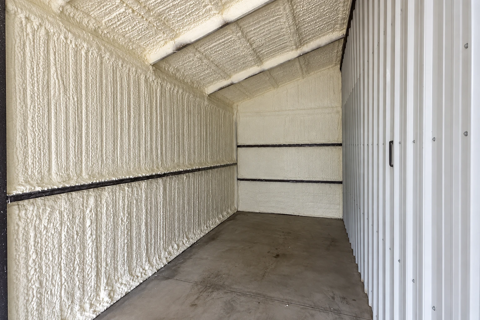 Narrow utility room with rough white spray foam covering the walls and sloped ceiling beside a metal-paneled wall.