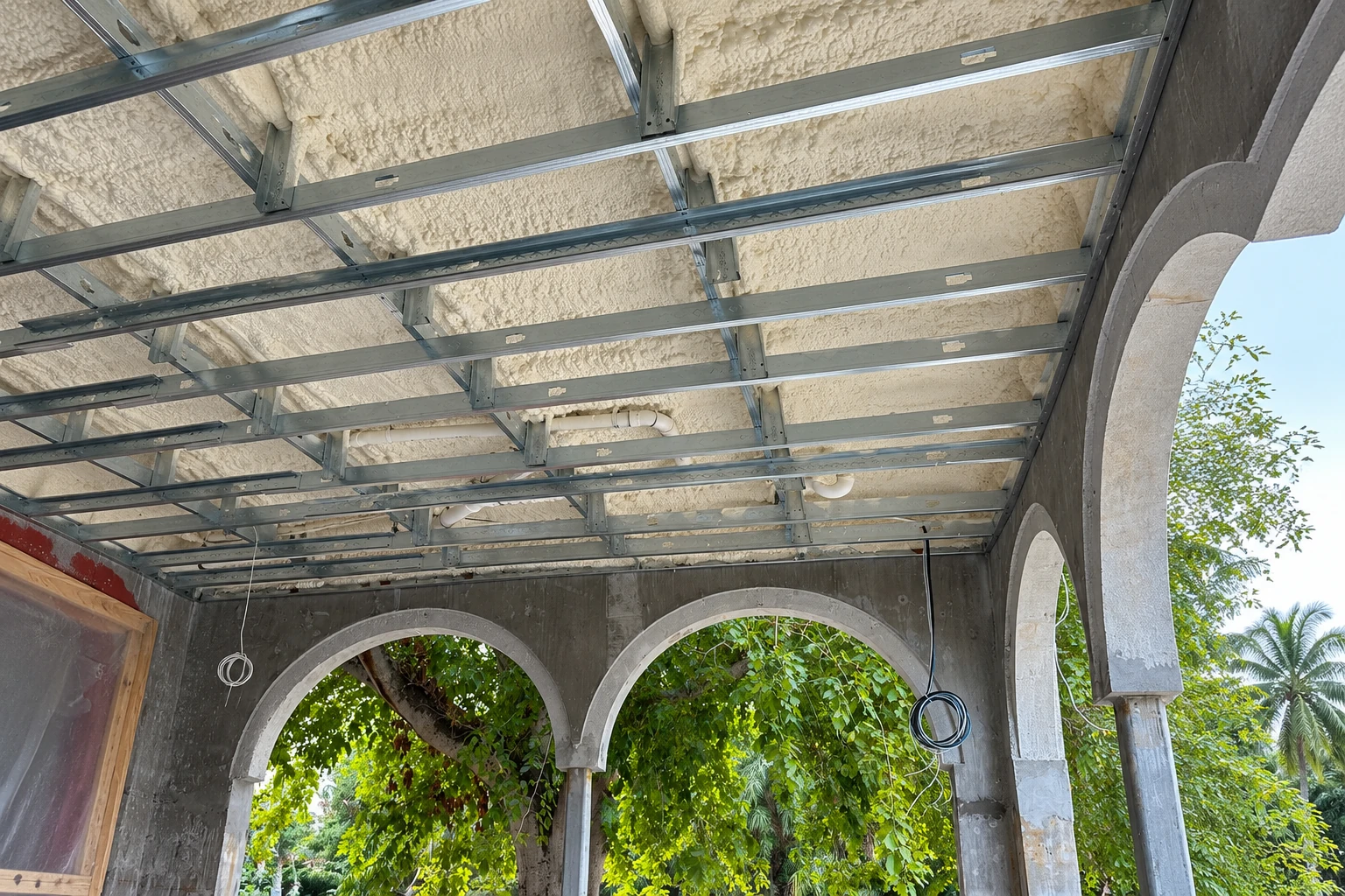 Spray foam coats the underside of a commercial ceiling, with metal framing and concrete arches below.