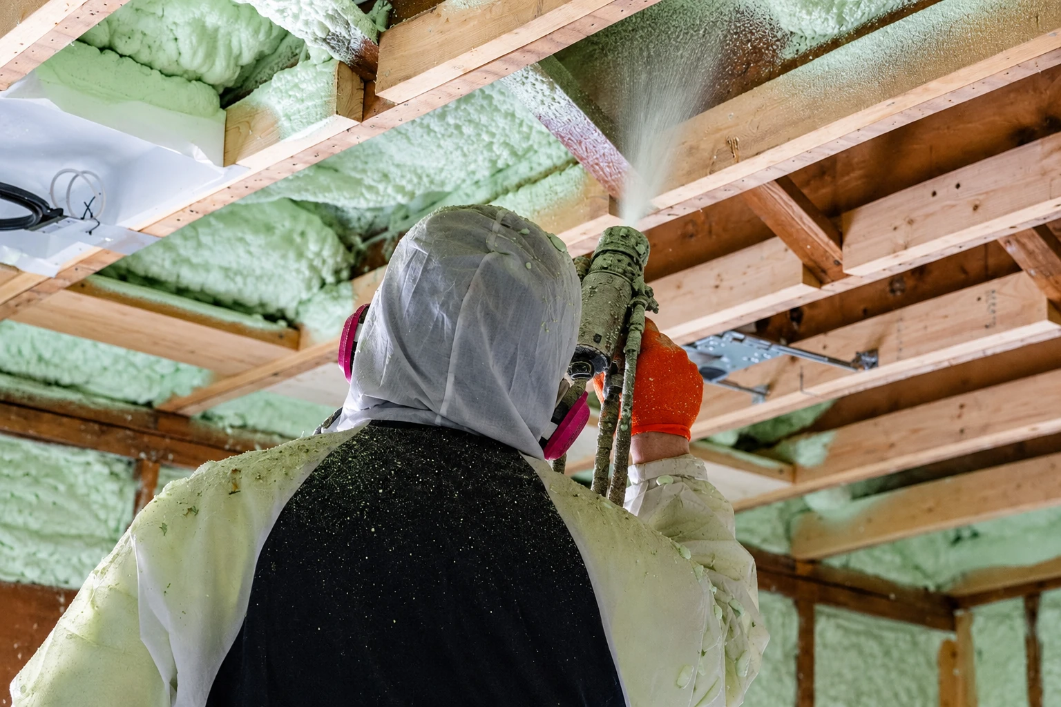A worker in protective gear sprays pale green open-cell foam between exposed wooden roof rafters.