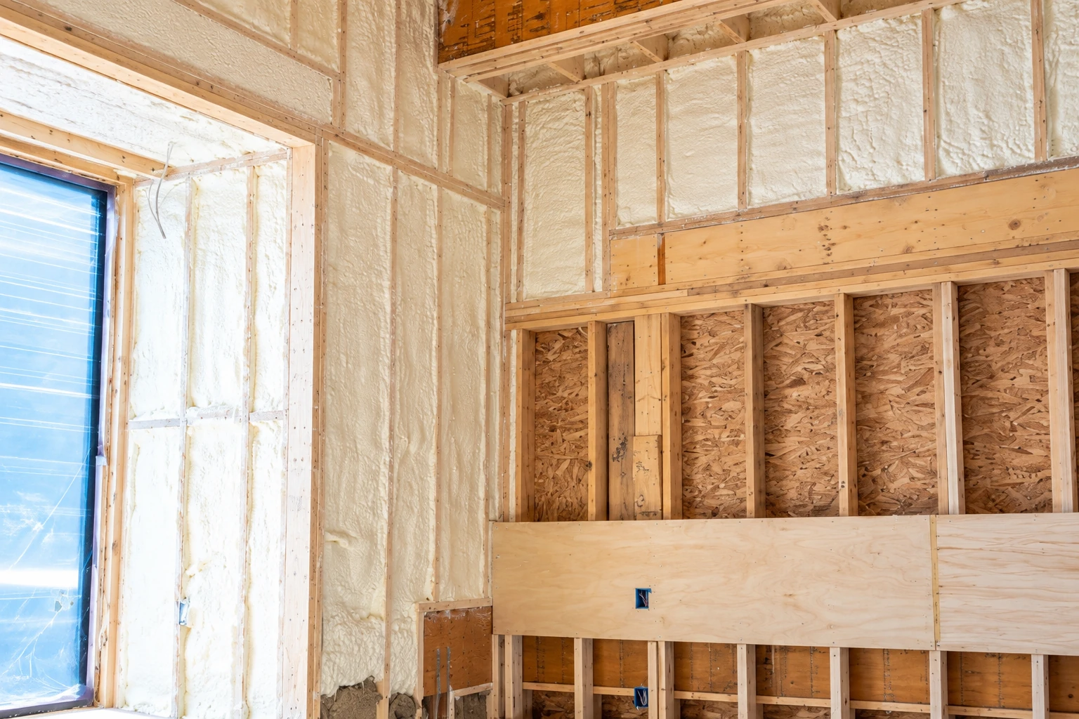 Unfinished wall framing with white spray foam insulation in the upper and left sections and exposed sheathing below.